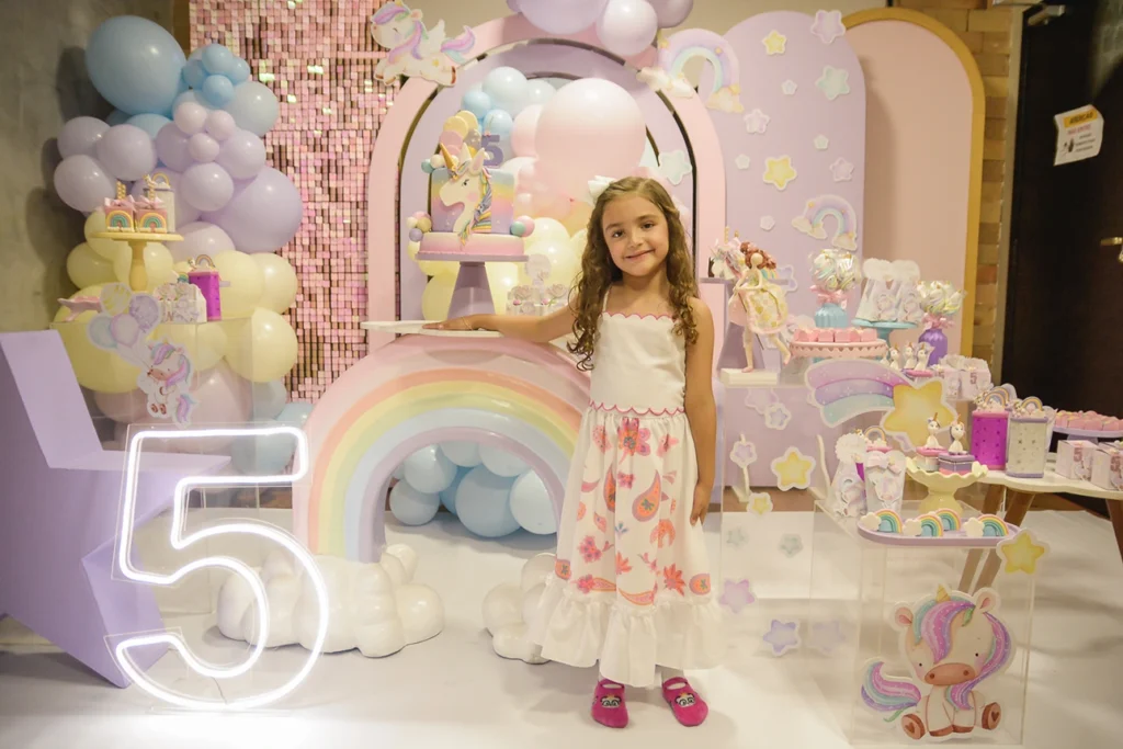 Foto de uma criança posando para foto em seu aniversário de 5 anos na qual o tema foi de unicornio. Ambiente nas cores predominante rosa e roxo.